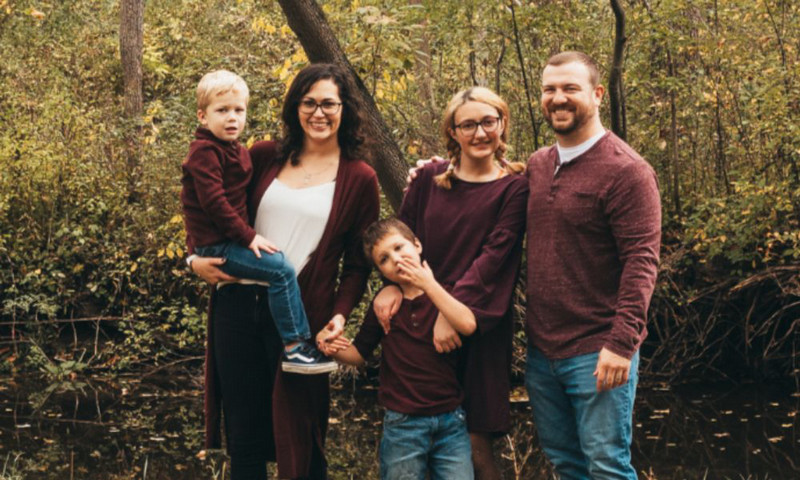 Family photo of Cassie Weis, husband Nicholas, and three children standing in front of green trees.