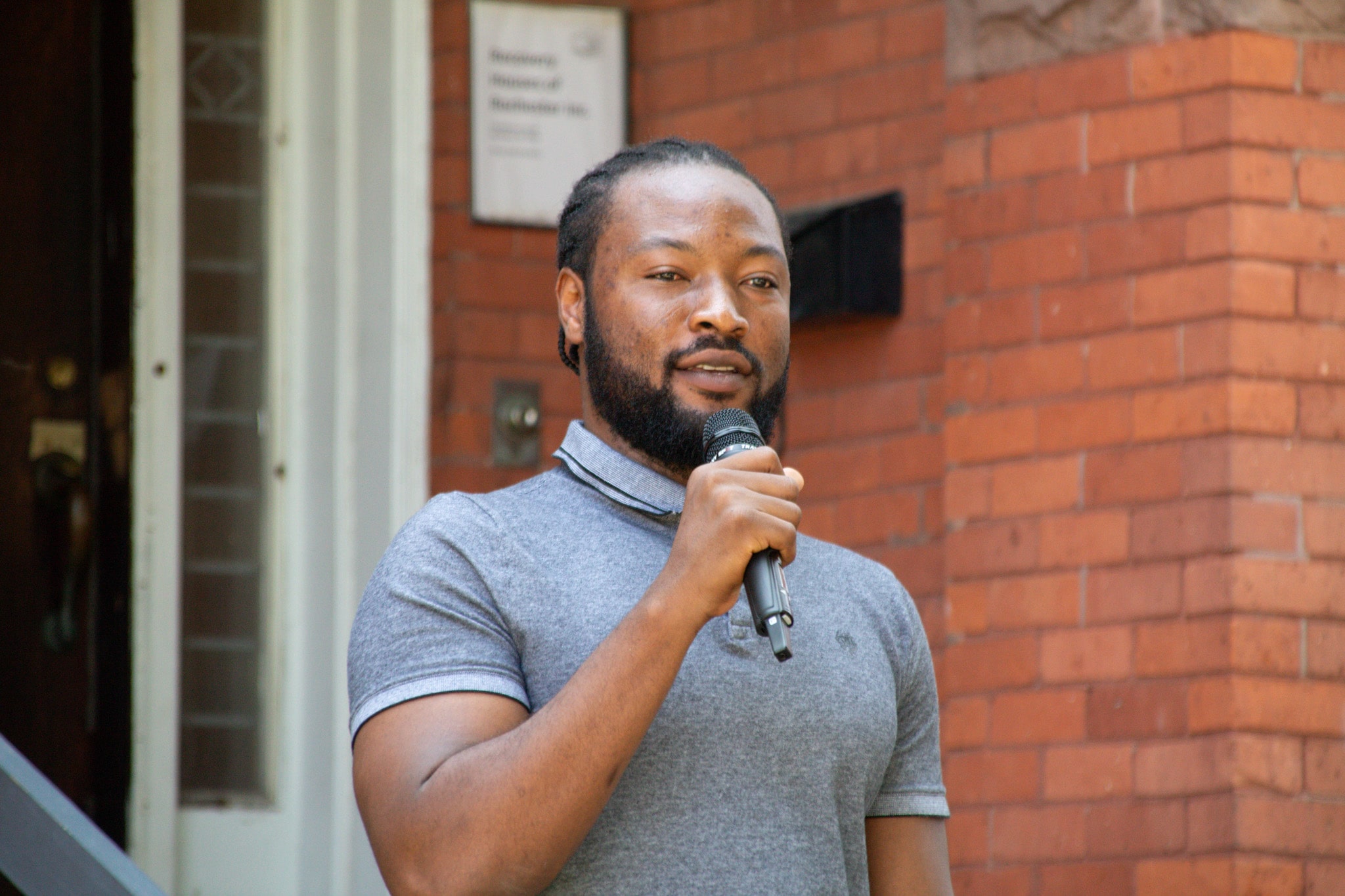 Gamji Rabiu Abu-Ba'are speaks at the Recovery Houses collaboration launch, standing on the house's front steps.