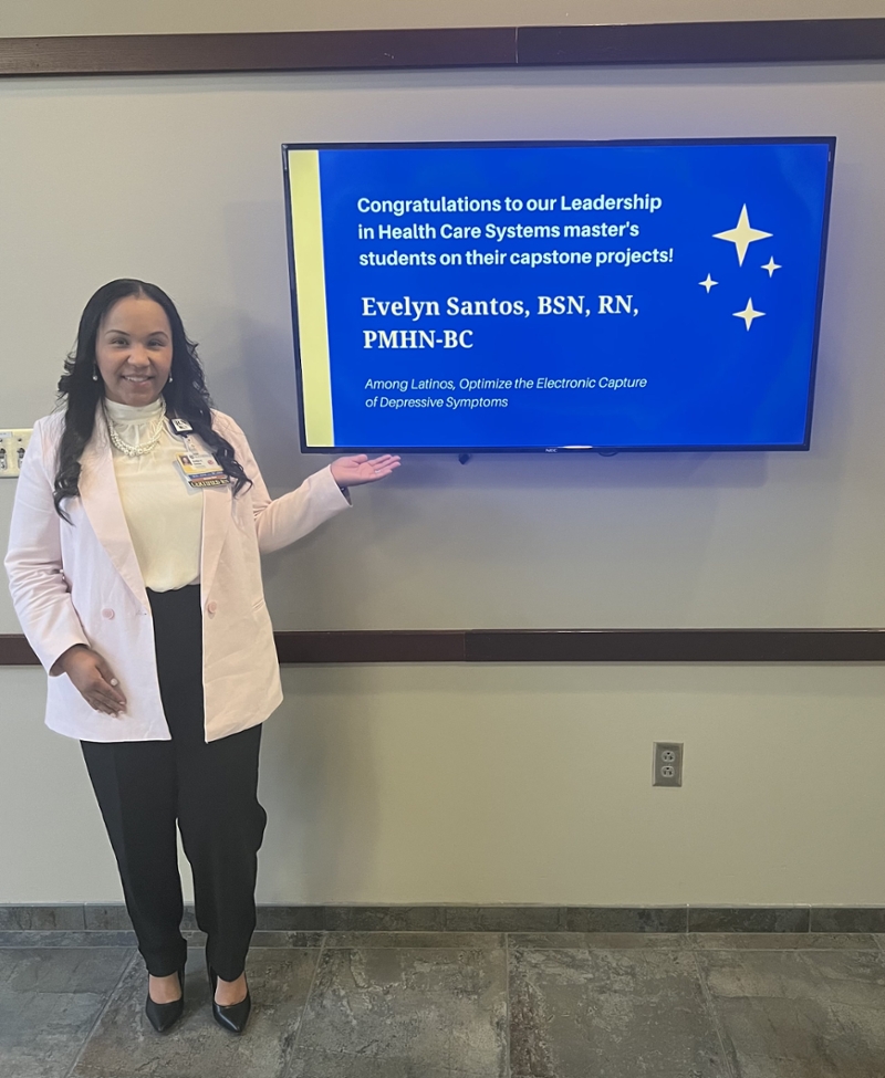 Evelyn stands next to a TV screen display congratulating LHCS students on their capstone presentations at the School of Nursing.