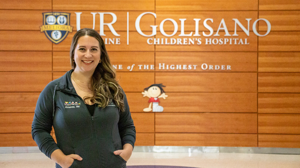 Amanda Welter stands in front of a wooden sign for Golisano Children's Hospital in a hallway.