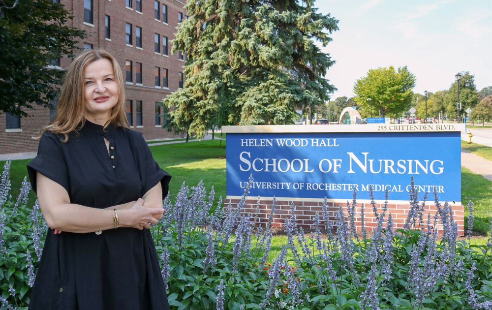 Brana smiles, wearing a black dress, standing next to the School of Nursing sign and purple flowers surrounding it.