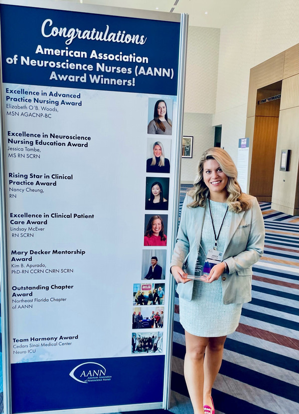 Jessica Tambe stands next to a banner congratulating the American Association of Neuroscience Nurses' 2024 award winners, while holding a glass trophy.