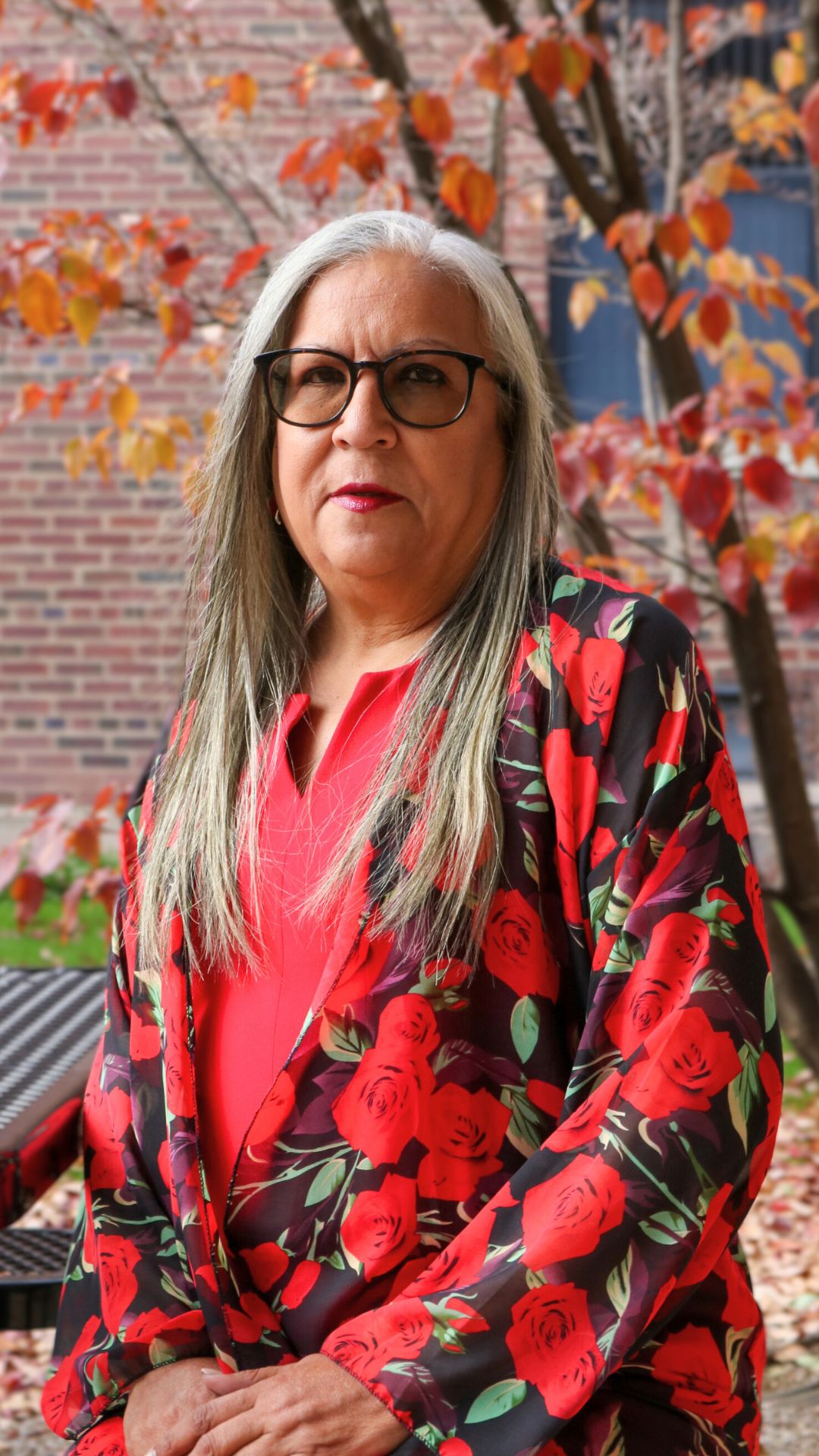 Portrait of Ivette Alvarez sitting outside at a table with orange autumn leaves behind her.